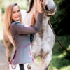 Schöne Frau mit Schimmel Pferd und modernem Turnierjacket in Grau